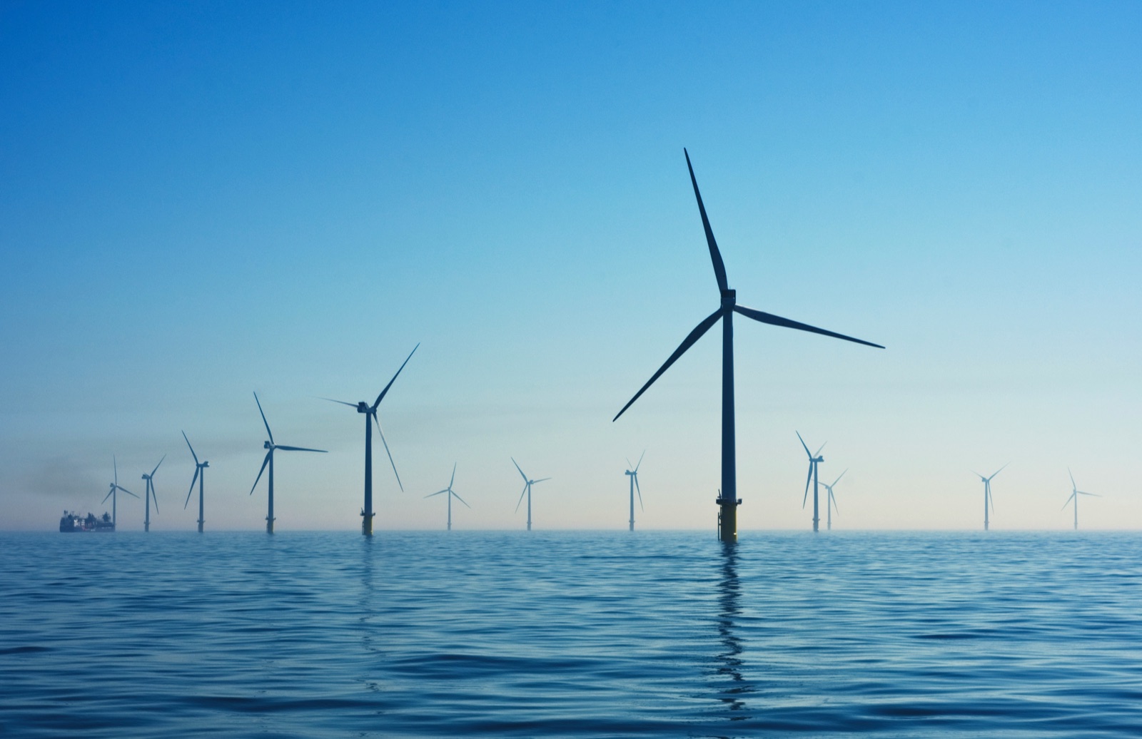 Offshore wind turbines rising from calm blue water beneath a hazy sky.