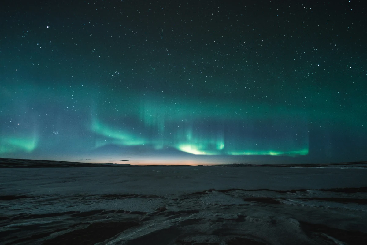 A green aurora stretches across a star-filled sky above a snowy landscape.