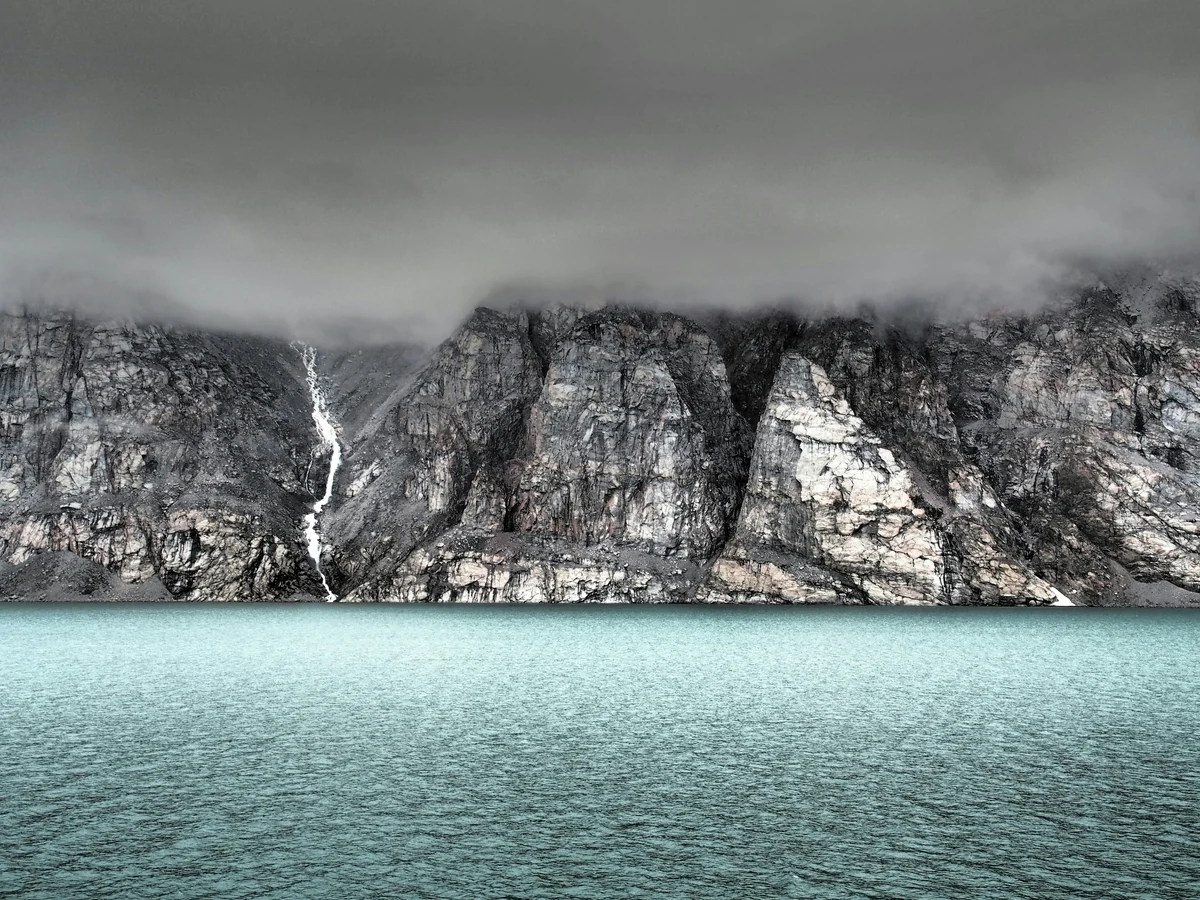 Low cloud hangs over steep rocky cliffs and a narrow waterfall above turquoise water.