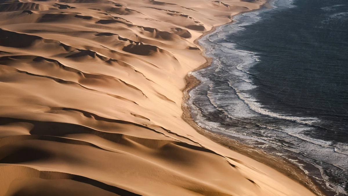 An aerial view of golden sand dunes meeting breaking waves and dark ocean water.