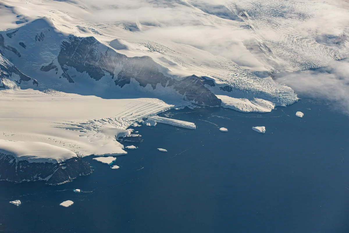 Aerial view of snow-covered coastal mountains and glaciers meeting dark blue water dotted with icebergs.