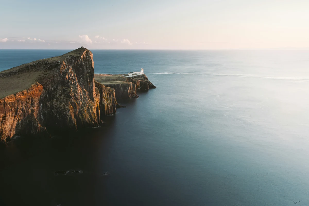 A lighthouse stands on a rocky headland above calm ocean water in low sunlight.