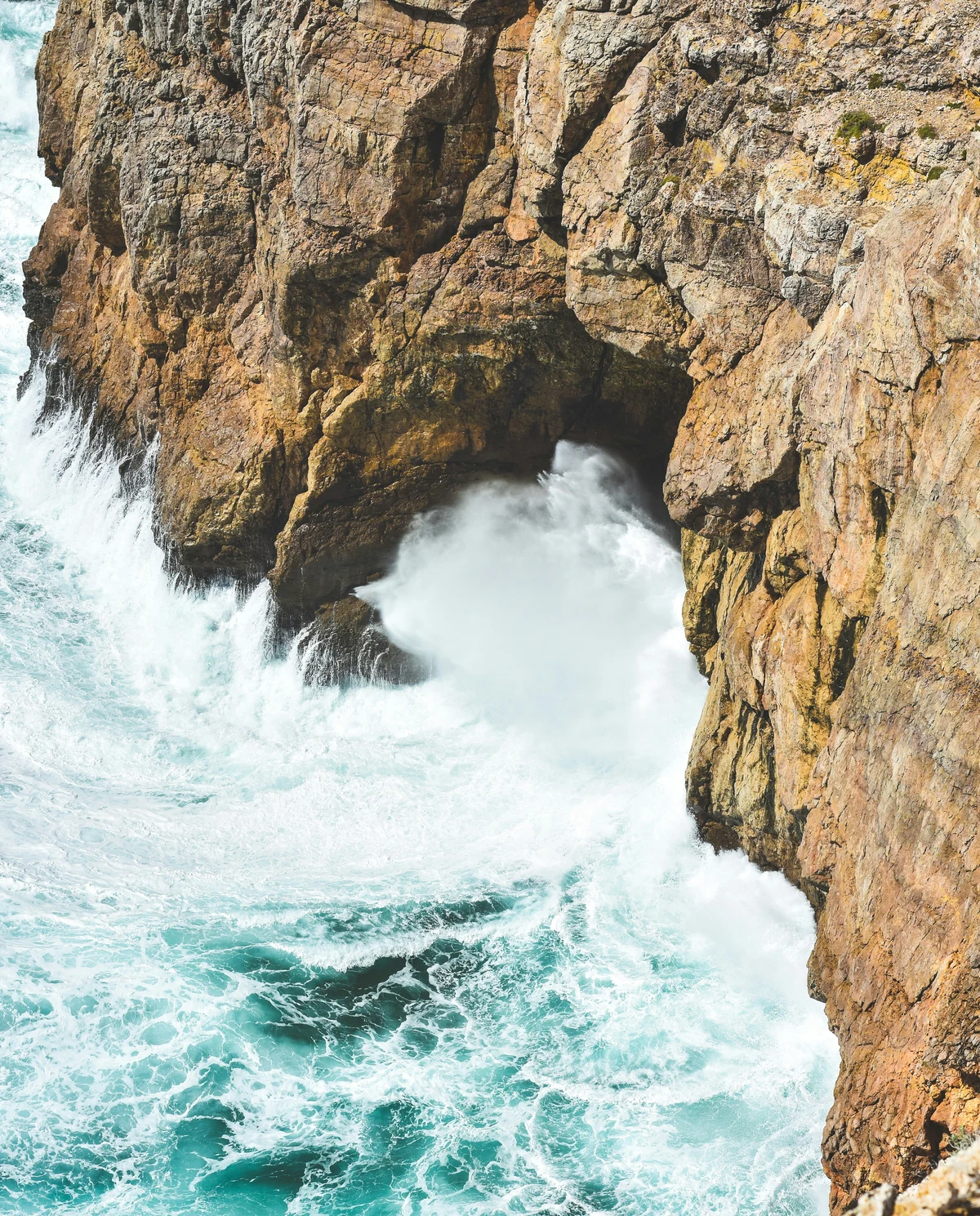 Foaming turquoise waves crash against steep, golden-brown coastal cliffs.